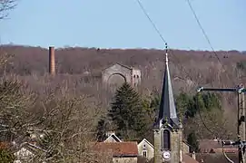 Trois bâtiment en ruines et une cheminée tronquée dépassent des arbres au-dessus de la collines surplombant le village.