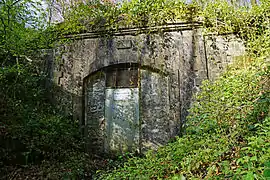 Entrée en pierre fermée par une petite porte métallique noyée dans la végétation.
