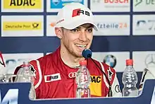 Photographie d'un homme en rouge et blanc, avec une casquette, de face, parlant à un micro, et souriant.