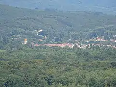 Clocher et toitures de maison émargeant d'une vaste plaine boisée et vallonnée.