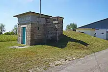 Un petit bâtiment cubique en pierre devant une butte de terre.