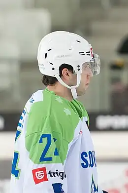 Photographie couleur d’un hockeyeur, de profil, de buste, vêtu d’un maillot blanc avec les épaules vertes