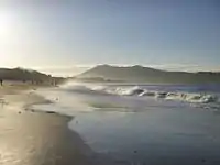 Photo en couleur d'une longue plage avec des vagues d'écume et des montagnes au loin, dans la lumière du matin.