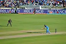 Photographie d'un match de cricket. Sous les yeux de défenseurs, un joueur cherche à frapper la balle alors qu’elle est en train de rebondir au sol.