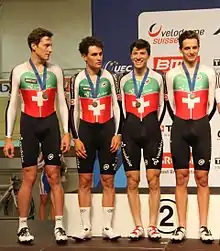 4 jeunes hommes debout, en pied, en tenue de cycliste, une médaille autour du cou