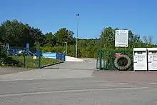 Photo couleur d'une rue route avec en arrière plan l'entrée grillagée d'une déchèterie aux bennes bleu