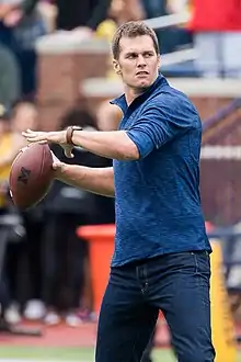 Tom Brady, en jean et veste bleue, lançant un ballon de football américain.