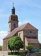 Vue arrière d'une grande église en grès rose surmontée d'un clocher comtois et d'un carillon.