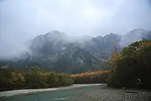 Photographie couleur d'une montagne entourée de nuage, avec à son pied une rivière.