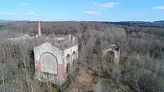 Photo aérienne hivernale montrant des bâtiments en ruine sans toiture et envahi par la végétation ainsi qu'une cheminée d'usine tronquée.