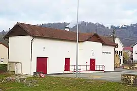 Bâtiment blanc disposant de plusieurs portes métalliques rouges, surmonté d'une petite cheminée d'où s’échappe une fumée banche.