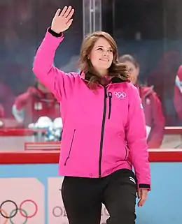 Photographie d’une femme, saluant de la main droite, portant une veste rose avec les anneaux olympiques.