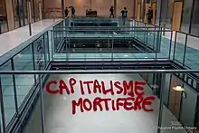 Tag à la peinture rouge : "Capitalisme mortifère" dans un hall propre et vitré.