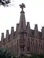 Un des angles avec le blason, les créneaux surmontés de barrettes doctorales, la croix de Gaudí, la colonne torsadée en briques, le T entre les créneaux et le monogramme JHS.