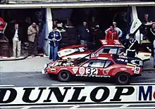 Photo d’époque d’une voiture de course rouge dans la ligne des stands du Mans