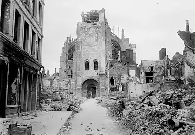 Photographie en noir et blanc. Tour-porche de la basilique avec ses parties hautes détruites. Au premier plan, nombreuses maisons en ruines.