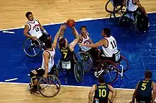 Photographie couleur de quatre joueurs de basketball en fauteuil roulant se disputent la balle.