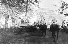 Une longue colonne de cavaliers avance le long d'une route. Un char est stationné à côté de la route