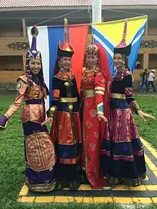 Quatre jeunes femmes en costumes aux couleurs vives posent devant les drapeaux de Russie et de Touva en prenant une posture gracieuse.