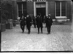 Quatre hommes marchent sur une cour pavée en chapeaux en parlant entre eux.