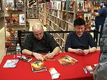 Deux hommes portant des lunettes, un caucasien et un asiatique, assis à une table et signant des bandes dessinées.
