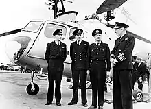 Photographie en noir et blanc du capitaine Burrell et de son équipage à côté d'un hélicoptère sur un navire.
