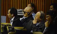 Ieng Sary est assis à la gauche de Nuon Chea et entourès de deux civils; ils portent des écouteurs sur les oreilles