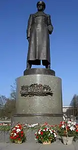 Photo en couleur d'une statue en pied sur piédestal, entourée de fleur.