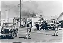 Trois soldats effectuant des signes de bras, devant deux voitures de l'armée et une rangée de petites maisons. En fond, on voit un nuage de fumée.