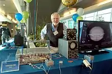 Photo d'une table avec dessus une machine et un oscilloscope, ainsi qu'un homme se tenant derrière.