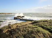 Au premier plan, rochers avec une toute petite jetée. En arrière-plan à gauche, le littoral du continent.