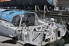 Vu de 3/4 arrière. L'arrière du bateau est blanc. On voit le tableau arrière, et on voit l'intérieur du cockpit, protégé par une casquette bleue.