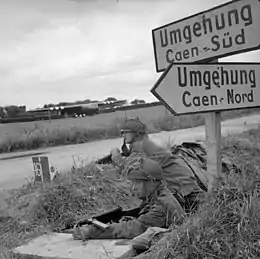 Photo noir et blanc de deux soldats en position près d'une route et sous un panneau routier.
