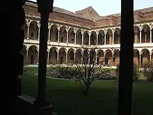 Image photographique d'un cloître. Deux colonnes au premier plan encadrent une cour verdoyante et un rond central avec au fond deux ailes à angle droit d'un édifice constitué s'arcades superposées sur deux niveaux surmontés par une toiture.