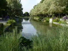 Photographie présentant un large canal aux rives bordées de massifs fleuris et de grands arbres.