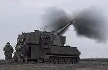 Canon automoteur en train de tirer. De la fumée sort du canon et trois hommes discutent à l'arrière du véhicule.