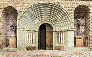La porte en bois dont le volet droit est ouvert est encadrée par des colonnades de pierres blanches soutenant un arc en plein cintre
