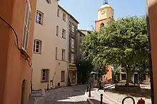 Une petite place à l'ombre d'un arbre, cernée de bâtiments de style provençal, dont une tour d'église.