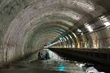 Intérieur d'un long tunnel de béton à la voûte courbe. À droite, une passerelle le longe. Au centre, une barque à moteur, avec trois personnes à son bord, avance sur l'eau sombre.