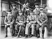 Photographie en noir et blanc représentant six hommes en uniforme militaire et coiffés de casquettes à visière. Deux d'entre eux sont debout et quatre assis