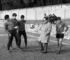 Photo en noir et blanc de 3 joueurs en survêtement qui jonglent, un homme en costume de ville et manteau, et un joueur en short.