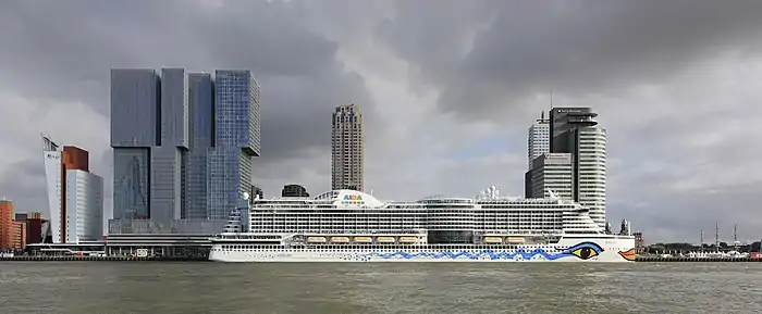 Accosté à un quai un grand bateau blanc, la coque avec une ligne de bleu. Derrière des immeubles modernes, ciel gris.
