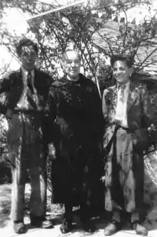 Photo noir et blanc de deux jeunes gens de part et d'autre d'une dame âgée, en pied, sous les ombrages