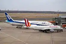 Photo de deux avions à la porte d'embarquement d'un aéroport. Au premier plan, un avion plus petit, aux couleurs blanches, oranges et rouges, cache la moitié d'un autre avion plus grand aux couleurs blanches et bleues, présent à ses côtés.