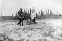 Deux hommes habillés de vêtements de fourrure posent devant un traîneau. Le sol est neigeux. À lʼarrière plan, une ligne dʼépinettes noires.