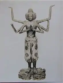 Front view of a standing statue with six arms and three faces. One pair of hands is joined in front of the body with the palms facing each other as if praying. A second set of arms is bend to the sides with the palms of the hands pointing upwards. Also the third pair of arms is bend somewhere between the positions of the other pairs of arms. The three faces point to the fron and both sides of the statue. The statue is embellished with sculpted and painted wide three-quarter length trousers and sandals.