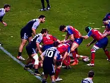 Dans un regroupement au sol de joueurs de rugby, l'un d'eux tend le bras pour présenter le ballon à son coéquipier.