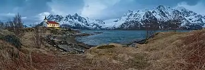 Une petite église s'élève sur une lande bordée par un bras de mer ; derrière, de hautes montagnes enneigées.