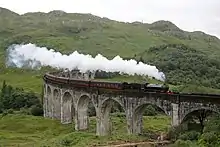 Dans un paysage de montagnes vertes, un train à vapeur rouge et noir traverse un pont en pierres avec des arcades.