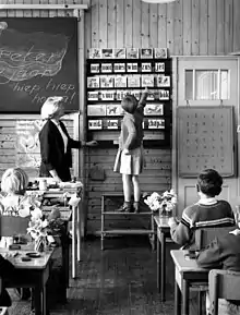 Une petite fille sur un escabeau désigne une image dans une salle de classe toute en bois.